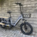 Image of a grey, long-tail cargo bike with tall handlebars and a basket in front, and a wooden deck and footrests by the cargo area.