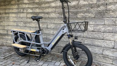 Image of a grey, long-tail cargo bike with tall handlebars and a basket in front, and a wooden deck and footrests by the cargo area.