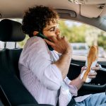 Young casually clothed man talking on a phone and eating sandwich while driving car