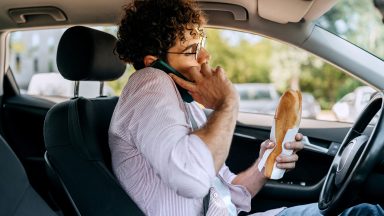 Young casually clothed man talking on a phone and eating sandwich while driving car