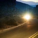 Blinding bright lights from a car pierce through the dark scene of a curved desert road at dusk. The lights form a star shaped glare. Double yellow lines on the paved road arc into the foreground. Mountains are visible in the distant background.