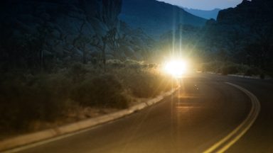 Blinding bright lights from a car pierce through the dark scene of a curved desert road at dusk. The lights form a star shaped glare. Double yellow lines on the paved road arc into the foreground. Mountains are visible in the distant background.