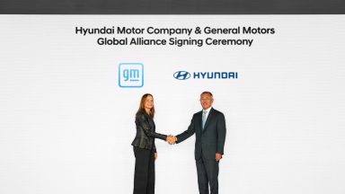 A woman and a man shake hands in front of a white background with GM and Hyundai logos above them