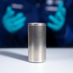 in the foreground, a large silver cylindrical battery cell. In the background, a pair of nitrile glove-clad hands