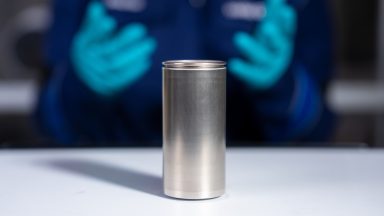 in the foreground, a large silver cylindrical battery cell. In the background, a pair of nitrile glove-clad hands