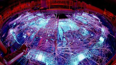 Image of a large, circular chamber covered filled with a lot of mechanical equipment, all of which is lit by blue internal glows and covered with massive, branching trails of lightning.