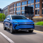 Front 7/8 view of a 2024 Chevrolet Equinox EV LT in Riptide Blue Metallic with a Summit White roof driving in a downtown area.