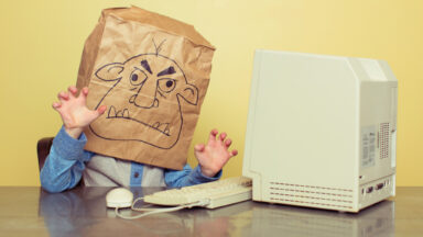 Picture of a person using an old Mac with a paper bag over his head. The bag has the face of a troll drawn on it.