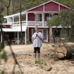 A woman looking at her smartphone in an area damaged by a hurricane. A house is in the background.