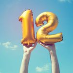 A gold foil number 12 balloon is held high in the air by male hands; photo taken outdoors on a bright sunny day, the sky is blue with some clouds. A vintage style effects has been added to the image.