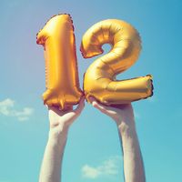 A gold foil number 12 balloon is held high in the air by male hands; photo taken outdoors on a bright sunny day, the sky is blue with some clouds. A vintage style effects has been added to the image.