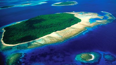 Aerial view of island, part of reef formtion, South Pacific, shows water on all sides, smaller islands in background and one in foreground.