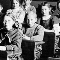 Vintage image of students at desks in a classroom with a teacher standing in the background. (education, learning)