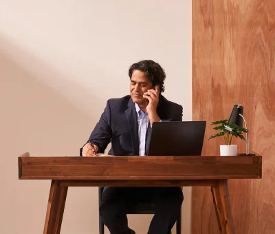 A man in a business suit and collared shirt speaks on a cellphone in his office, while writing a note on the desk.