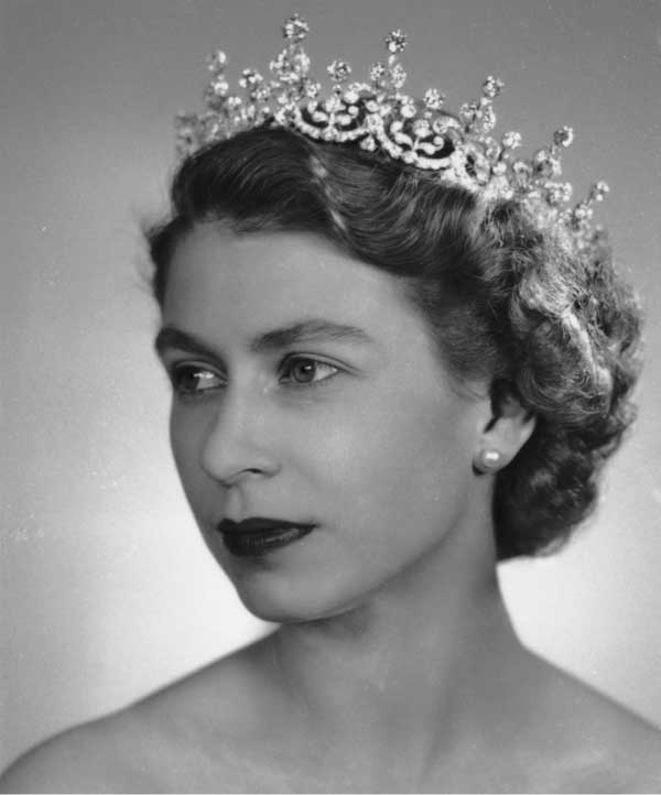 A tight crop, black and white photograph of Queen Elizabeth II from the shoulders up. She is looking to the side, wearing a diamond crown with dark lipstick.