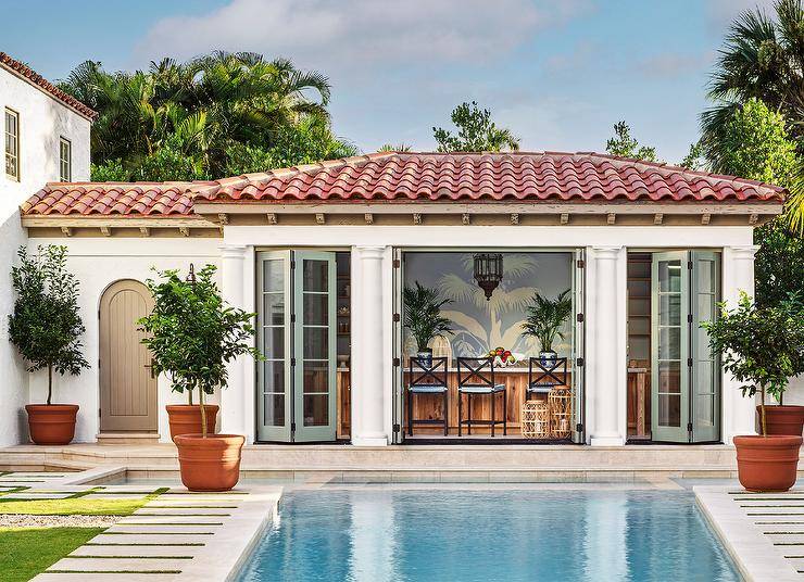 Pool house bar features green folding doors that open to an in ground rectangular pool and black stools with blue cushions.