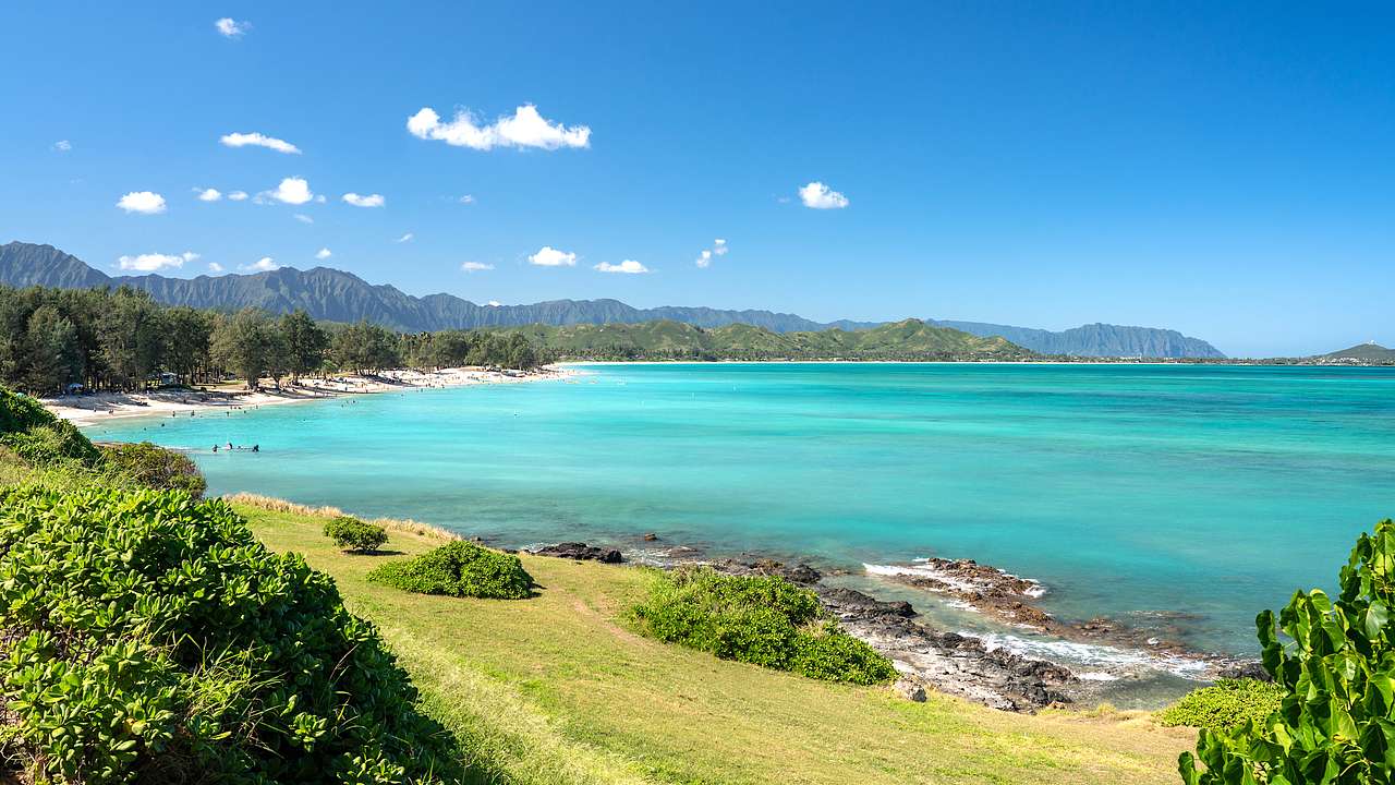 Side view of a sandy white beach with blue water next to a grassy incline with rocks