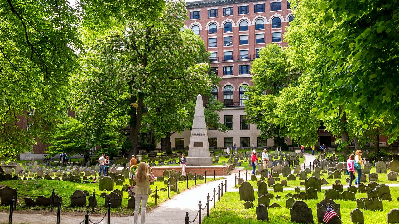 A path and tombstones are set on a lawn with trees and an obelisk in the center