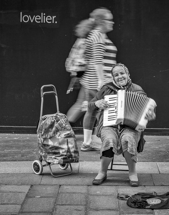 A woman playing accordion on the street with the text 'lovelier' above her head 