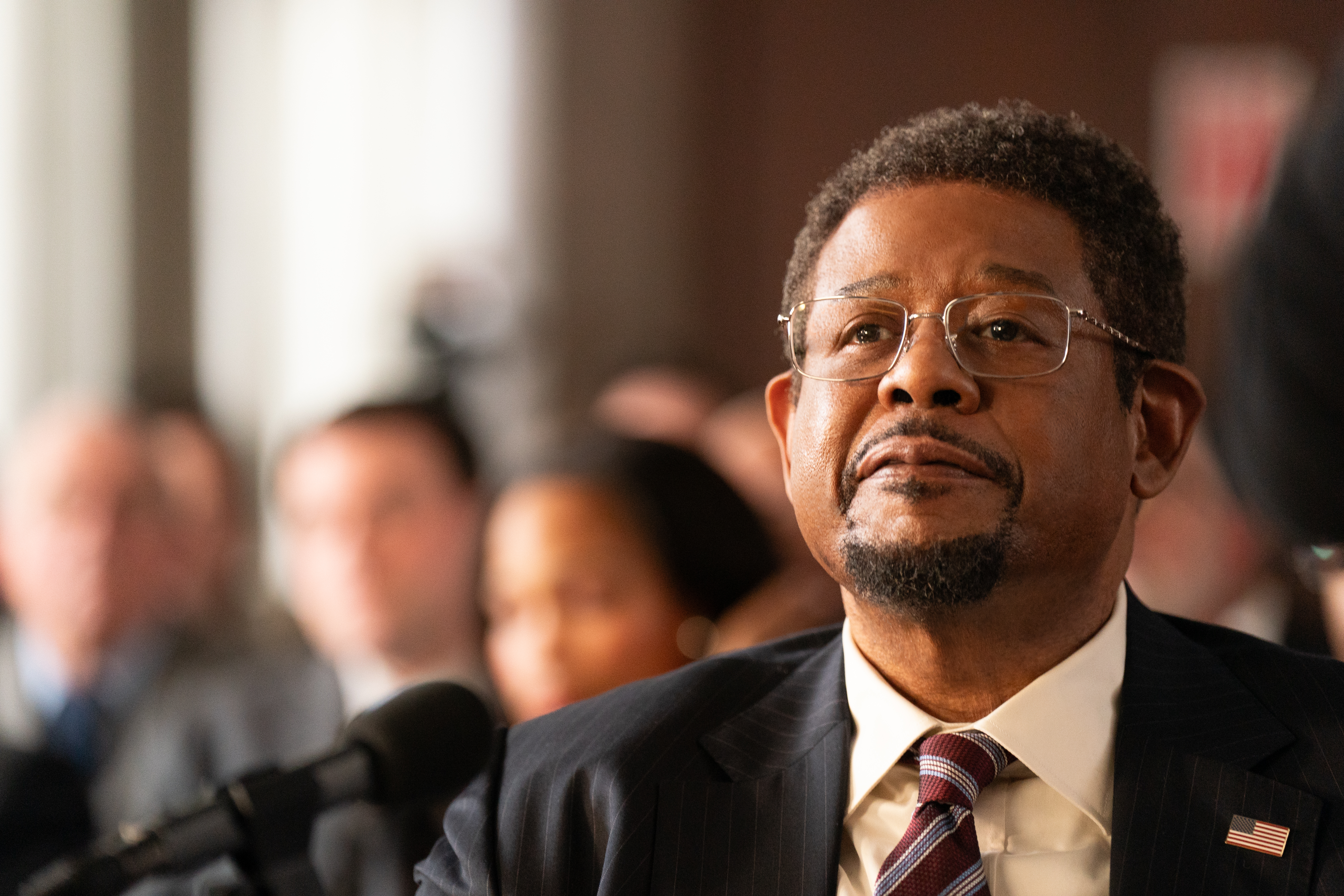 A Black man with glasses and a suit looks out straight ahead in front of a microphone.