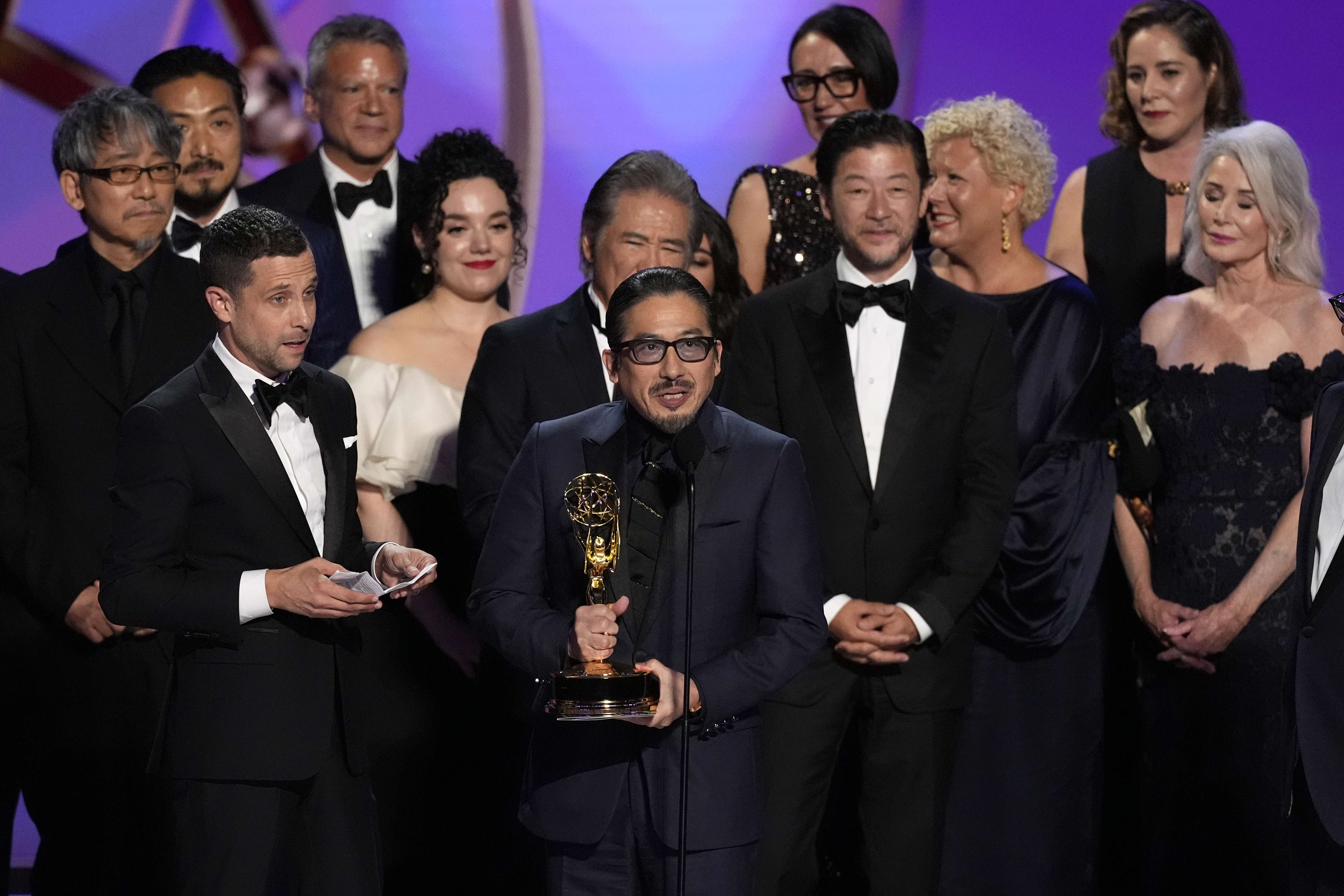 A group of people in suits and gowns stands on a stage behind a man holding a golden award statuette. 