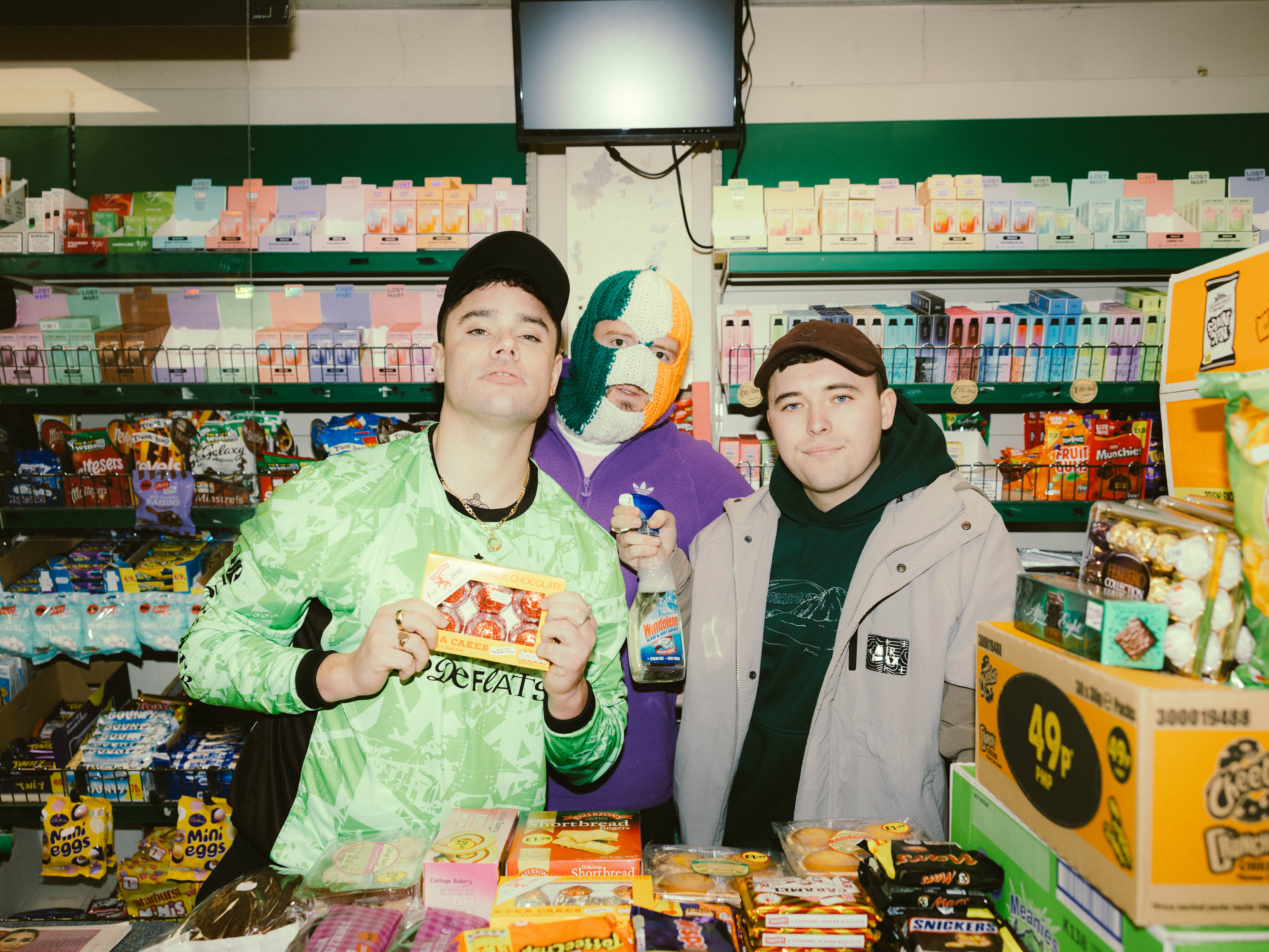 Moglai Bap, DJ Provai, and Mo Chara from Kneecap. They are standing behind the counter at a convenience store, holding various goods. Moglai Bap is wearing a green long-sleeve shirt. DJ Provai is wearing an Irish flag colored balaclava and a purple sweater. Mo Chara is wearing a black hoodie and a light brown duster. 