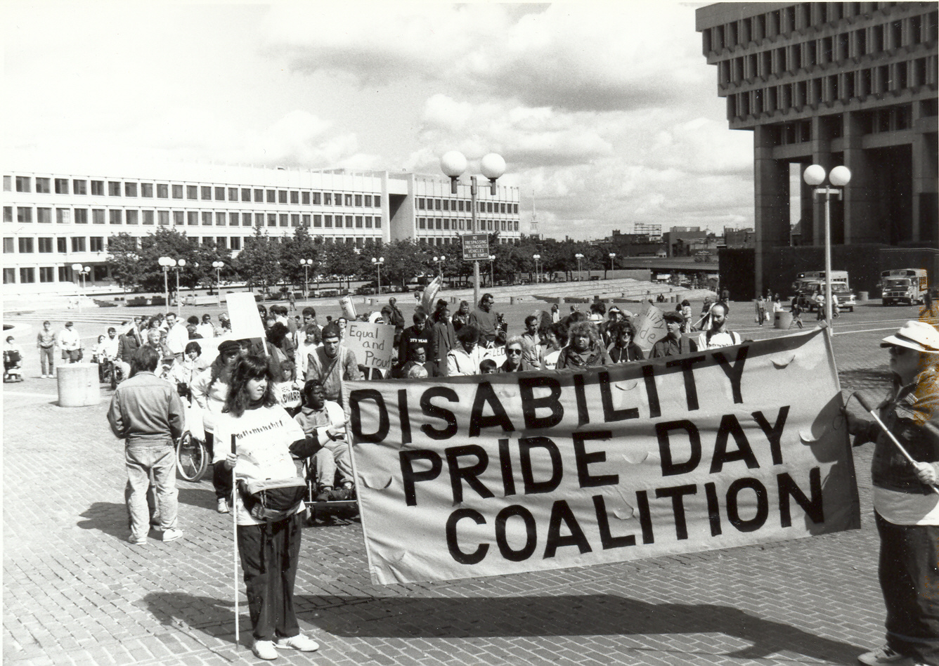 A black and white photo. A group of people, some in wheelchairs and holding white canes gather on City Hall Plaza. At the front, they hold a banner that says 'Disability Pride Day Coalition'