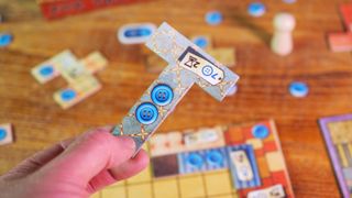 A hand holding a playing piece from Patchwork, with game components on a wooden table in the background