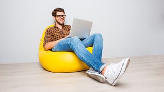 A cheerful-looking man sitting on a beanbag chair and working on a laptop.