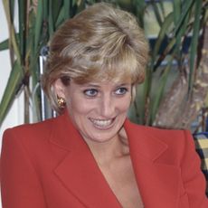 Princess Diana laughs while wearing a red two-piece suit by Catherine Walker while visiting London Lighthouse for people affected by HIV and AIDS on October 8, 1996, and Camilla Parker Bowles wears a black sleeveless dress with a pearl necklace