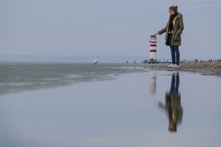 A woman in a scarf and jacket stands on the right and holds out a hand to touch the top of a lighthouse, which is actually behind her in the distancetop of a smaller lighthouse on a beach.