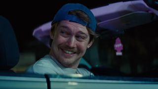 Joe Alwyn sitting in a car, smiling and looking behind him.