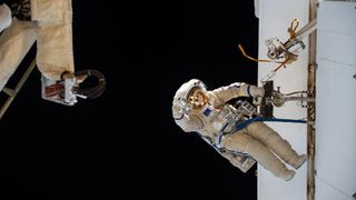 an astronaut in a white space suit hangs on to a metal bar on the outside of a large white structure. the blackness of space fills the background