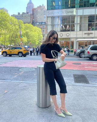 Woman in NYC with black capris, a black shirt, mules, and a sage green bag