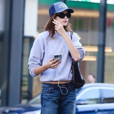 Kaia Gerber wears a blue hat, black sunglasses, wired headphones, blue jeans, a black shoulder bag, and a light blue crewneck sweatshirt.