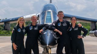 Four people in dark jumpsuits lean on a jet.