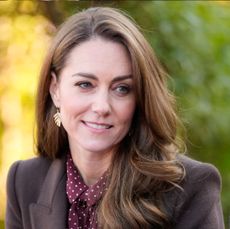 Kate Middleton wearing a brown coat and maroon dress looking to the side and smiling with green and yellow leaves behind her