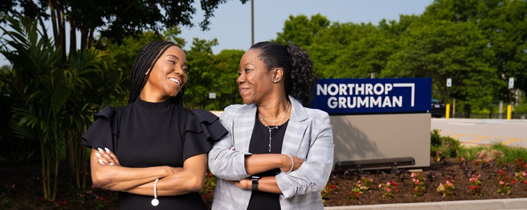 Two women in business professional dress and red heels stand, with arms crossed, in front of a Northrop Grumman facility.