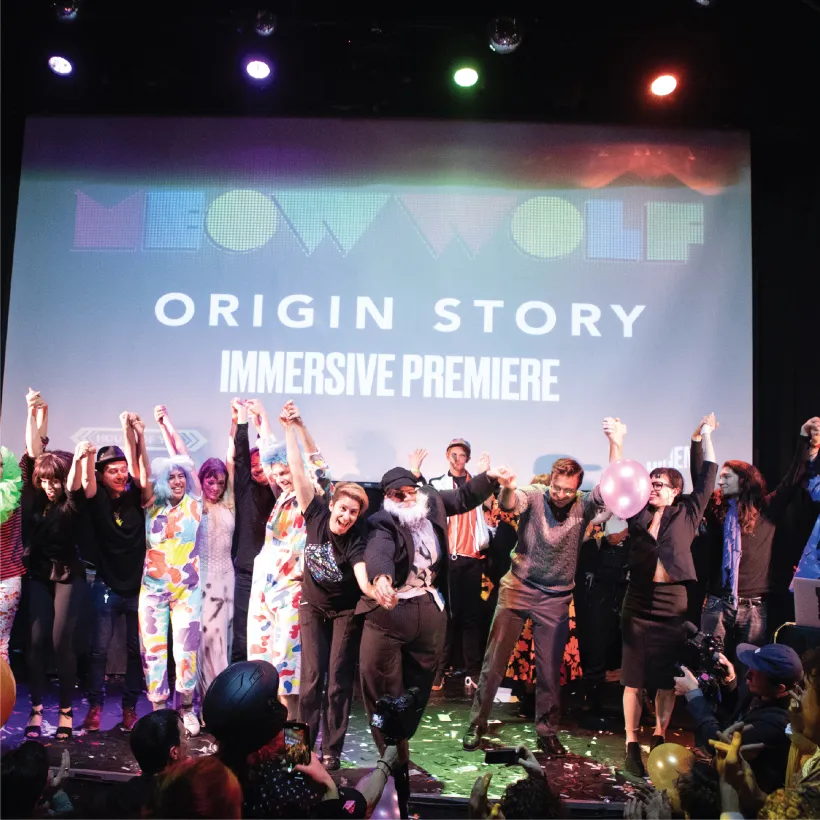 large group of people standing on a stage in front of a screen that says "Origin Story"