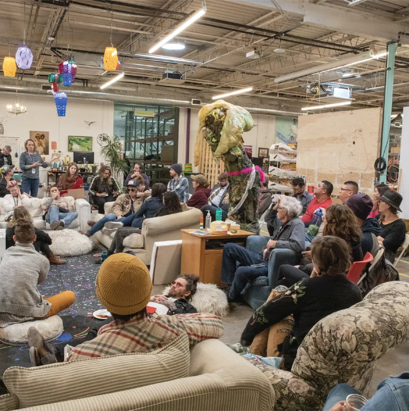 People sitting on different couches, bean bags, chairs, and the floor inside an art warehouse