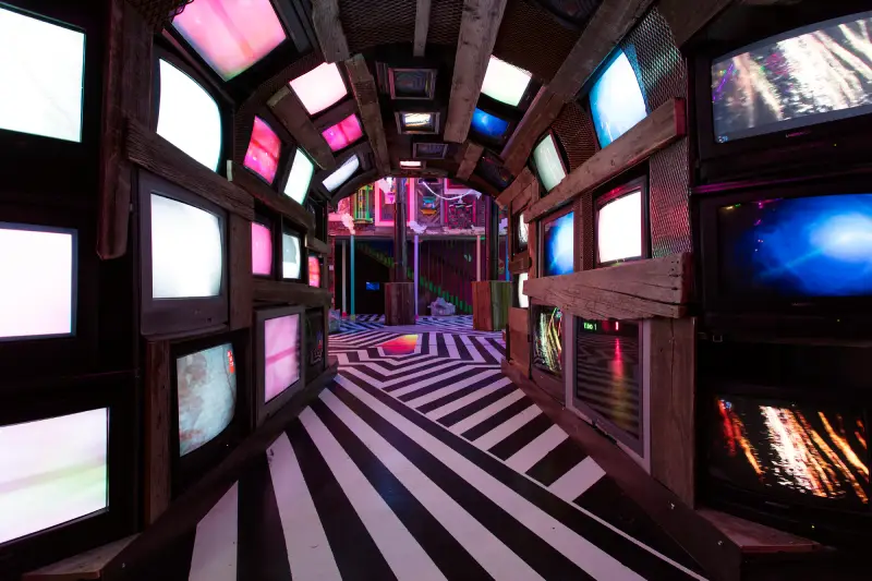 A tunnel made of tv screen walls with a black and white striped floor leads into another room at Meow Wolf Santa Fe.