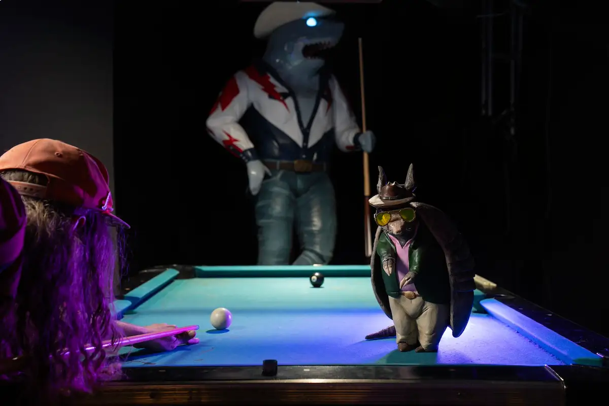 A person playing at a pool table with two creature cowboys. 