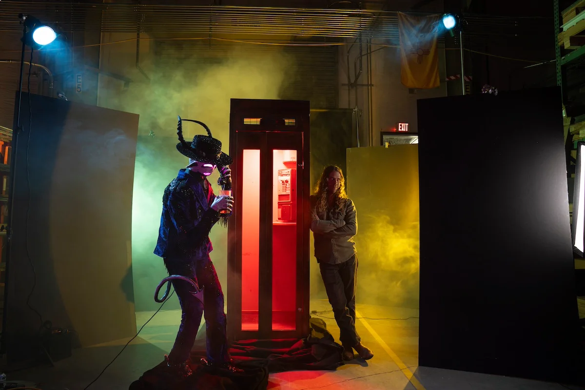 A person leaning against a telephone booth emitting red light, with a strange statue of a creature on the other side. 