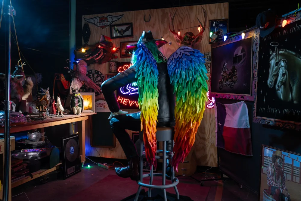 A cowboy sits on a chair facing away, with a pair of rainbow wings. 