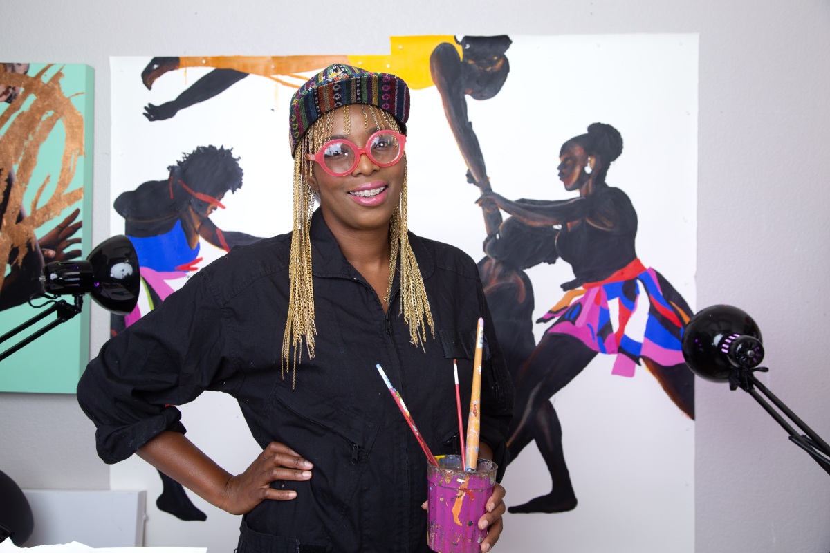 Artist Dawn Okoro wearing gold chains under a hat and red glasses, standing in front of her artwork holding a cup with paint brushes and smiling into the camera.