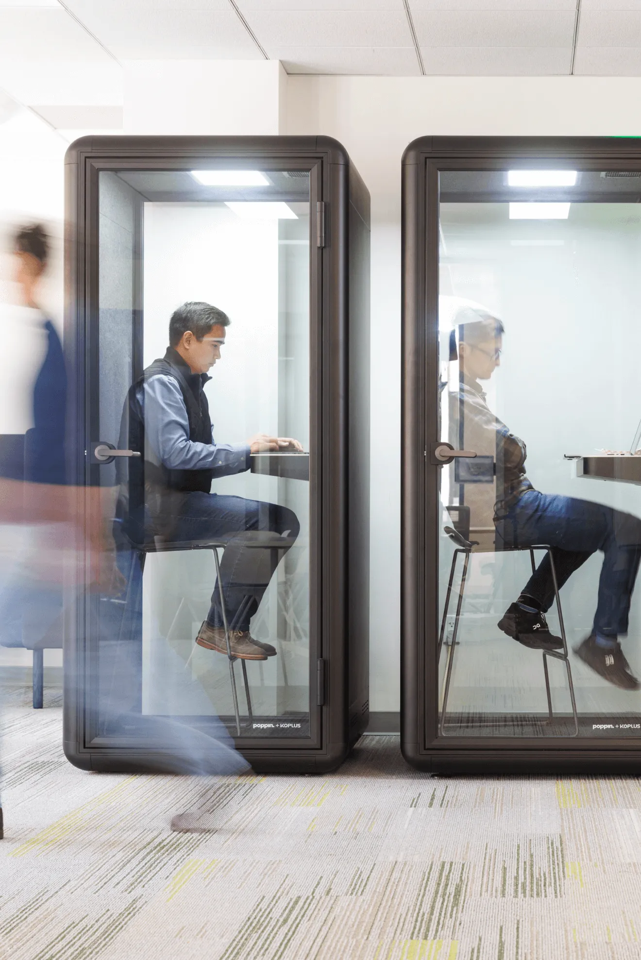 Two people sitting next to each other in clear phone booths in a co-working space
