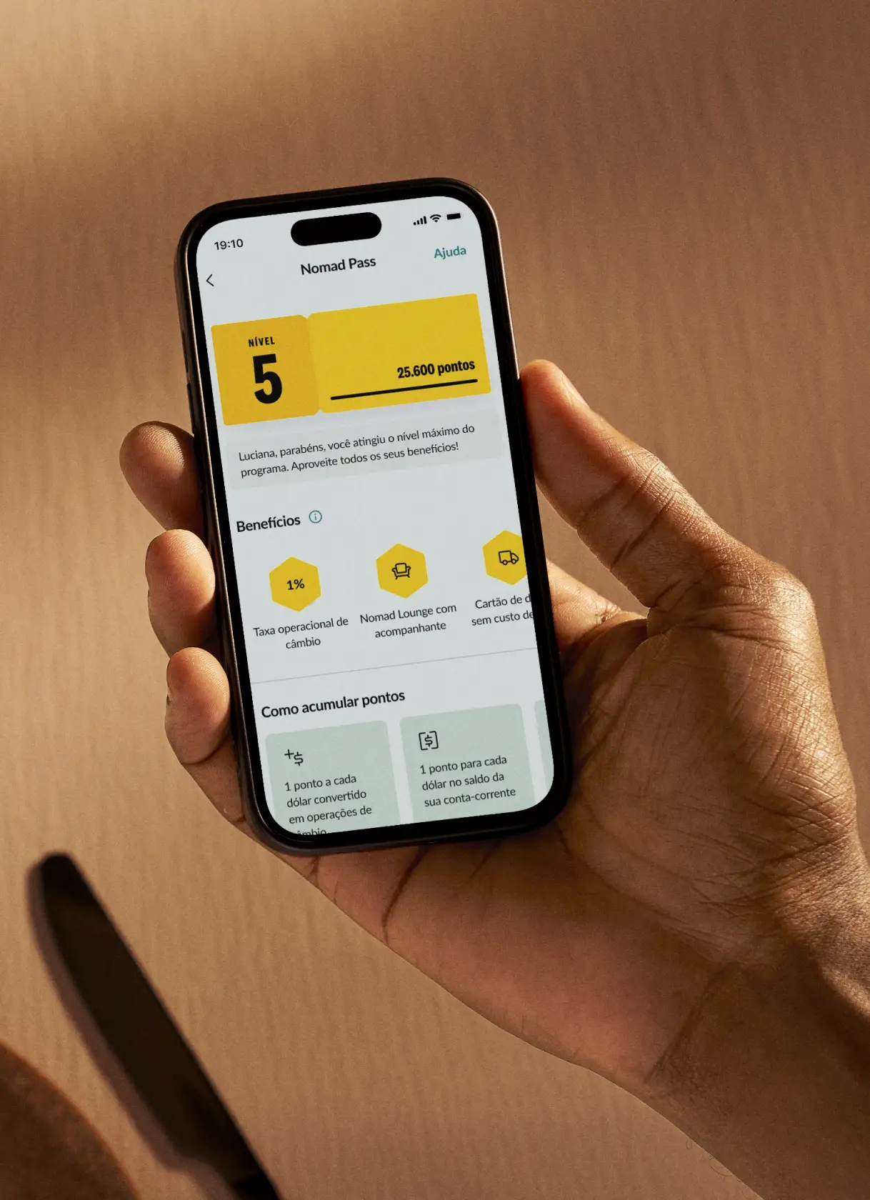 Uma mão segurando um smartphone exibindo a tela de um aplicativo chamado 'Nomad Pass'. A tela mostra o nível 5 com 25.600 pontos, benefícios como taxa operacional de câmbio, acesso ao Nomad Lounge com acompanhante e cartão de débito sem custo de emissão. Abaixo, há informações sobre como acumular pontos.