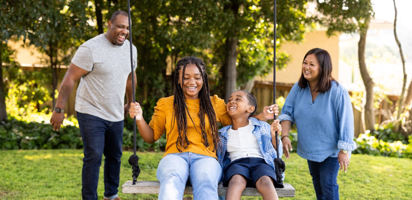 A happy family standing outside with two kids on a swing, and the parents on either side of them