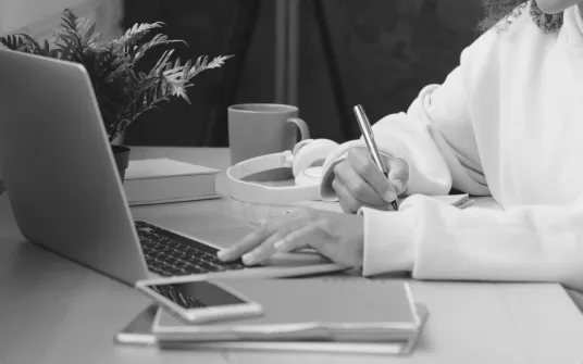 Black & white photo of a person doing research on their laptop while taking notes with pen & paper.