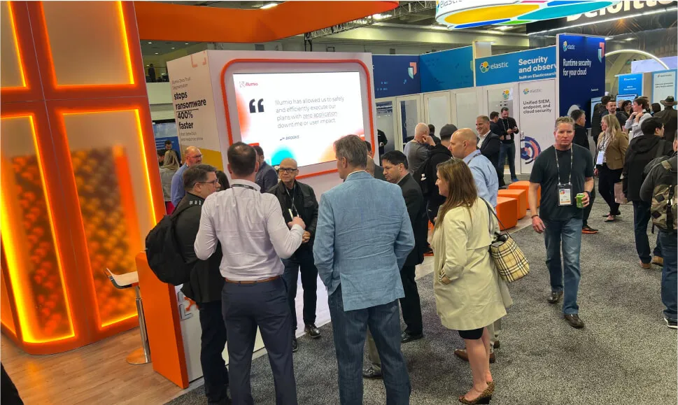 People gathered around an Illumio convention booth with a sign quoting the company 'Brooks' saying "Illumio has allowed us to safely and efficiently execute our plans with zero application downtime or user impact."